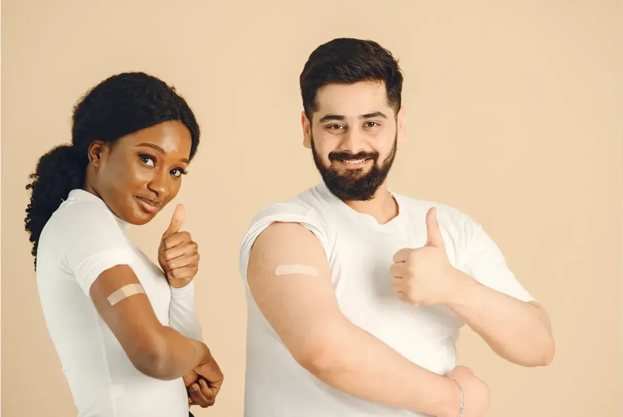 Two smiling adults giving a thumbs-up, showing bandages on their arms after getting vaccinated